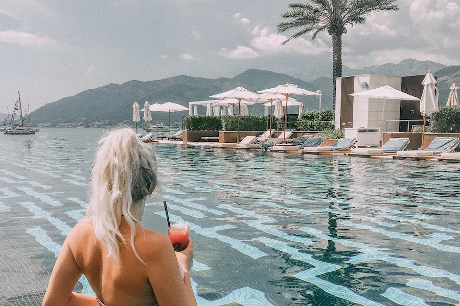 The Best thing to do in Montenegro - Porto Montenegro. Back of a blonde girl sitting in the blue pool at Porto Montenegro looking out over the water and mountains in the background
