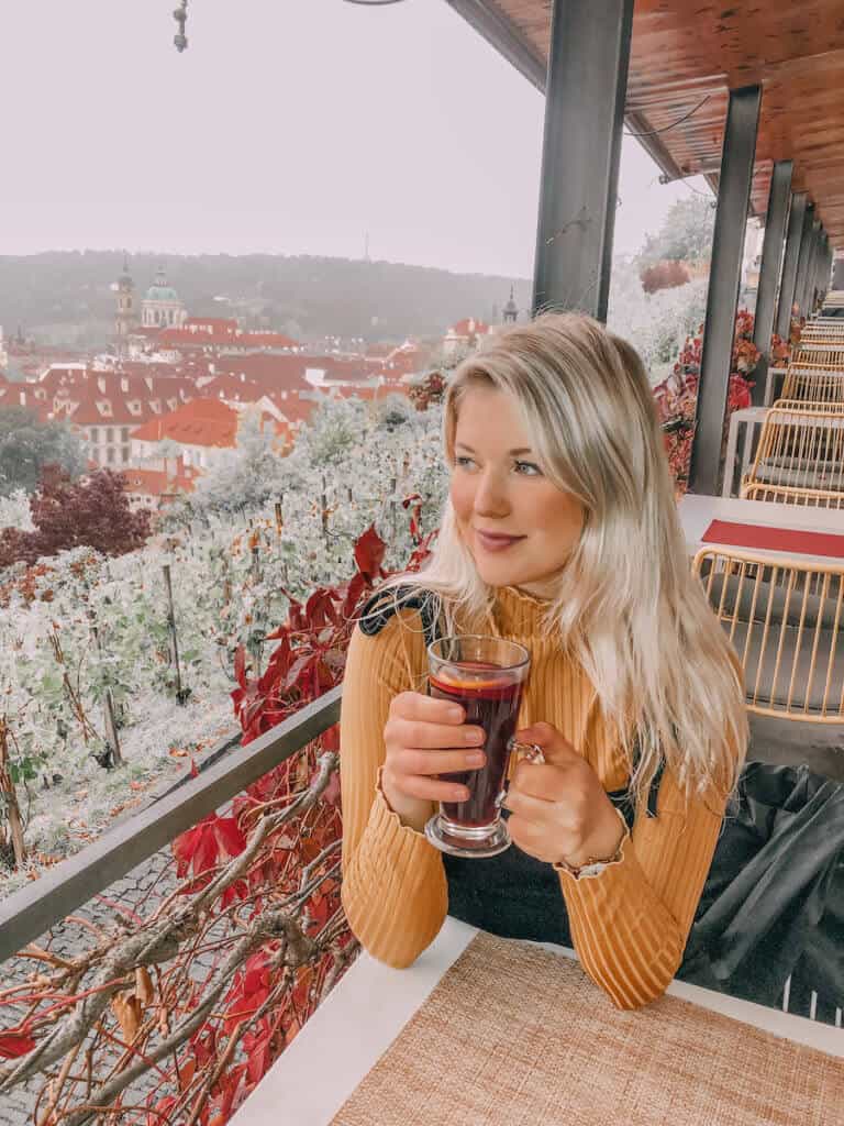 Blonde girl sitting on a terrace with a glass of wine and the city of Prague in the distance