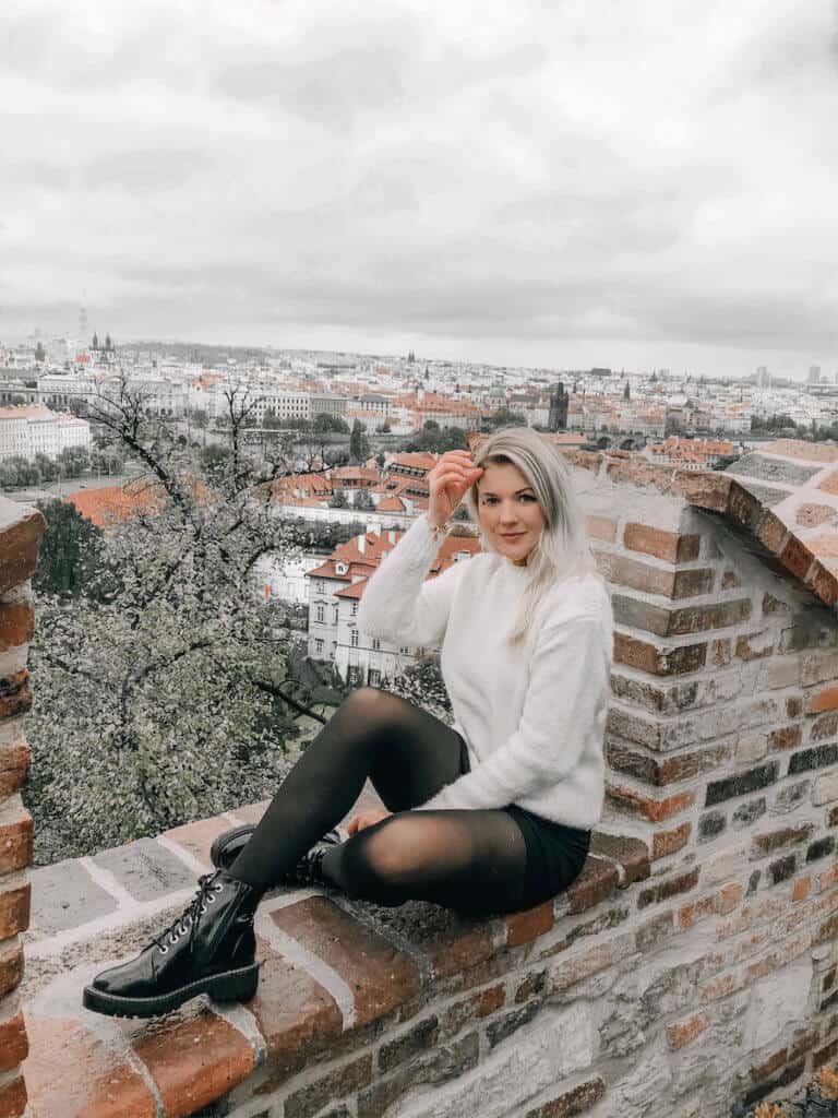 Blonde girl sitting on the edge of the Prague castle and the city of Prague in the distance