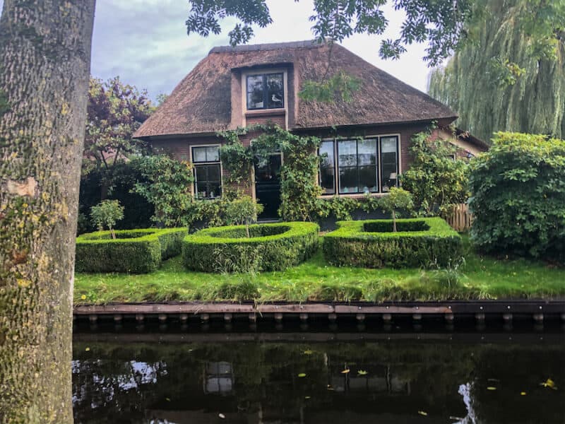 Thatched roof houses with greenery in front of it and a canal in front of that