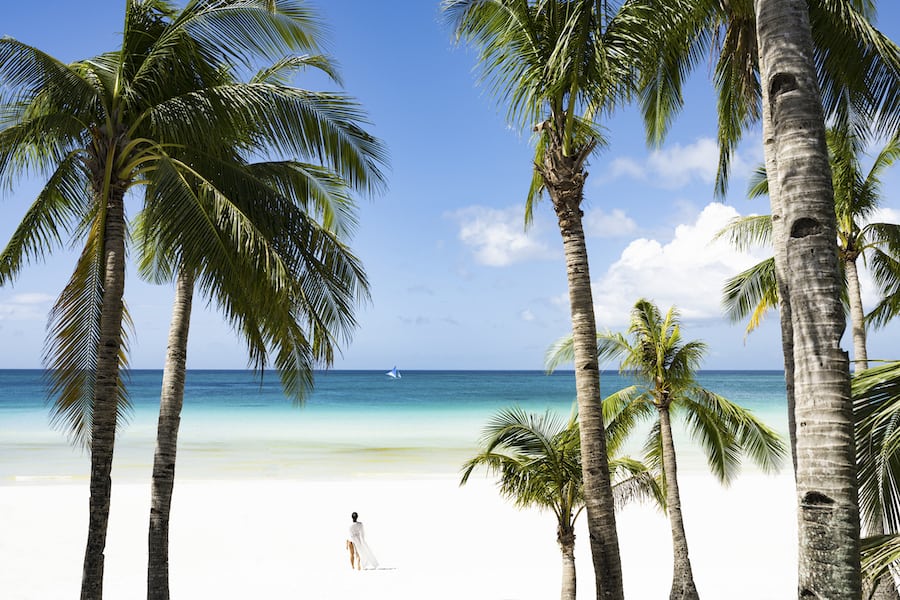 Stunning view of woman walking on a white sand beach bathed by a turquoise sea, beautiful coconut palm trees in the foreground. White Beach, Boracay Island, Philippines.
