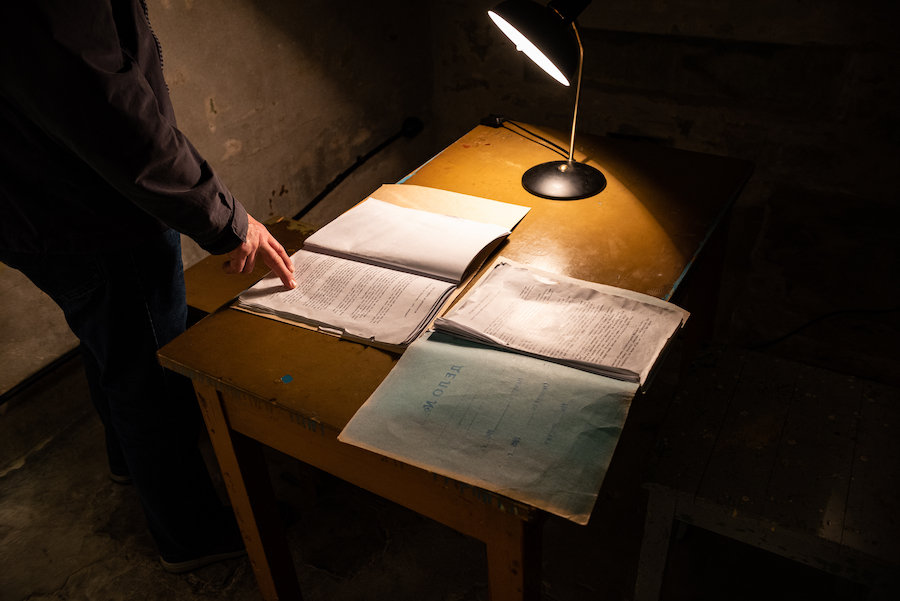 A dramatic, atmospheric, low-key, dark photo of a man's hand on a historic typed KGB document on a desk with desktop light in the KGB Museum in old town Tallinn.