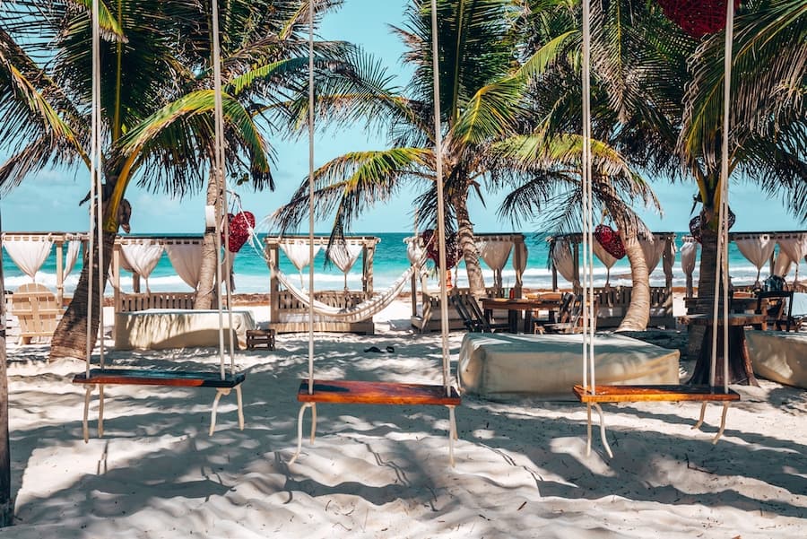 Hanging wooden swing seats tied up with rope on sand at beautiful beach resort with canopy, in tulum, Mexico.